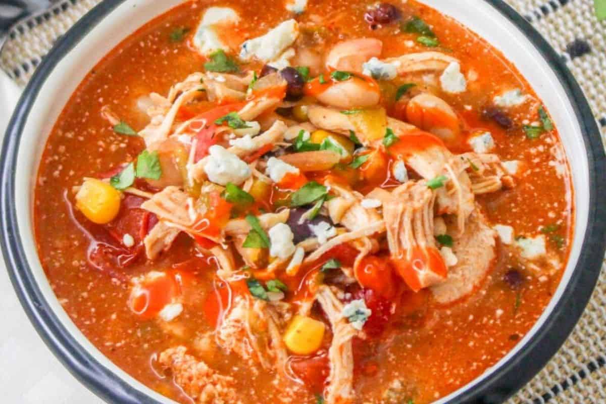 A bowl of Slow Cooker Buffalo Chicken Chili filled with shredded chicken, black and white beans, corn, diced tomatoes, and topped with blue cheese crumbles, chopped parsley, and a drizzle of buffalo sauce.