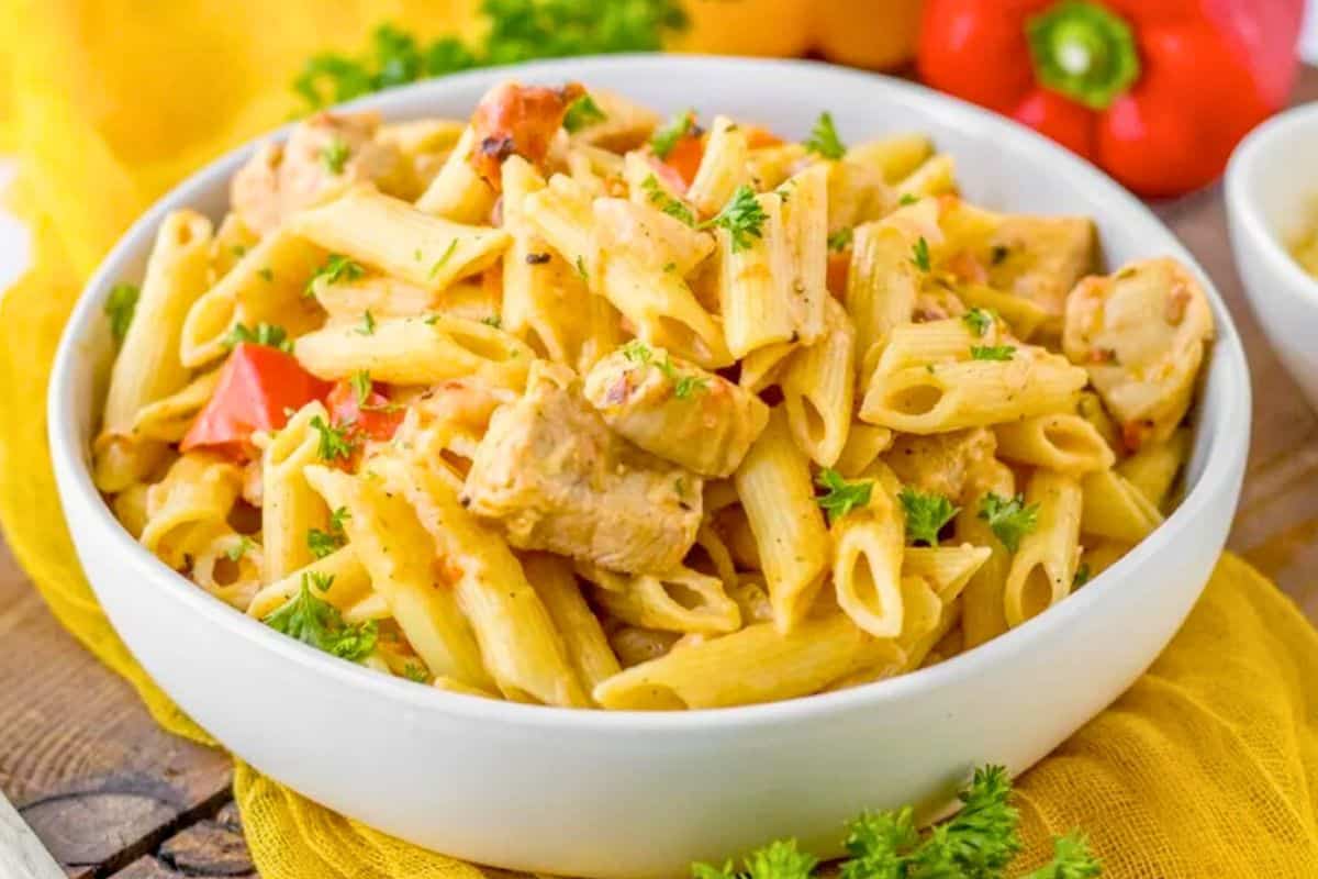 A white bowl filled with penne pasta, diced chicken, red bell peppers, and garnished with chopped parsley.