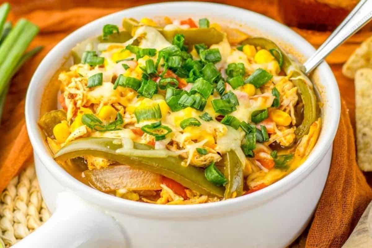 A white bowl filled with shredded chicken, corn, sliced green peppers, melted cheese, and chopped green onions, with a spoon inside.