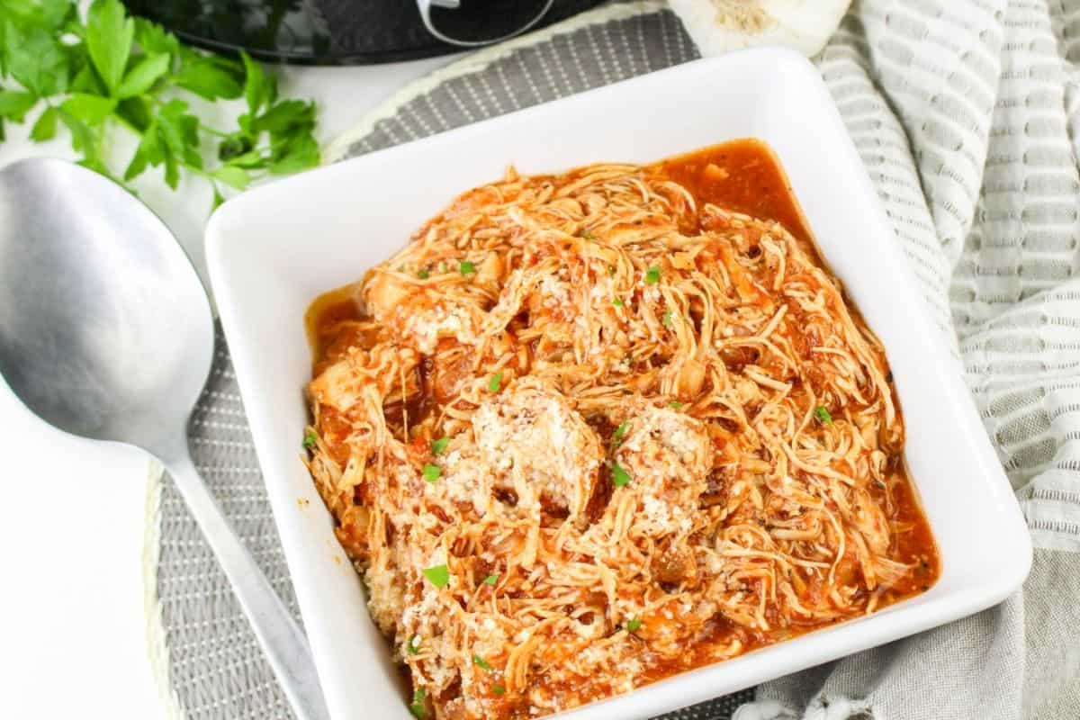A square white bowl filled with shredded chicken in tomato sauce, garnished with herbs, next to a silver spoon and a striped napkin.