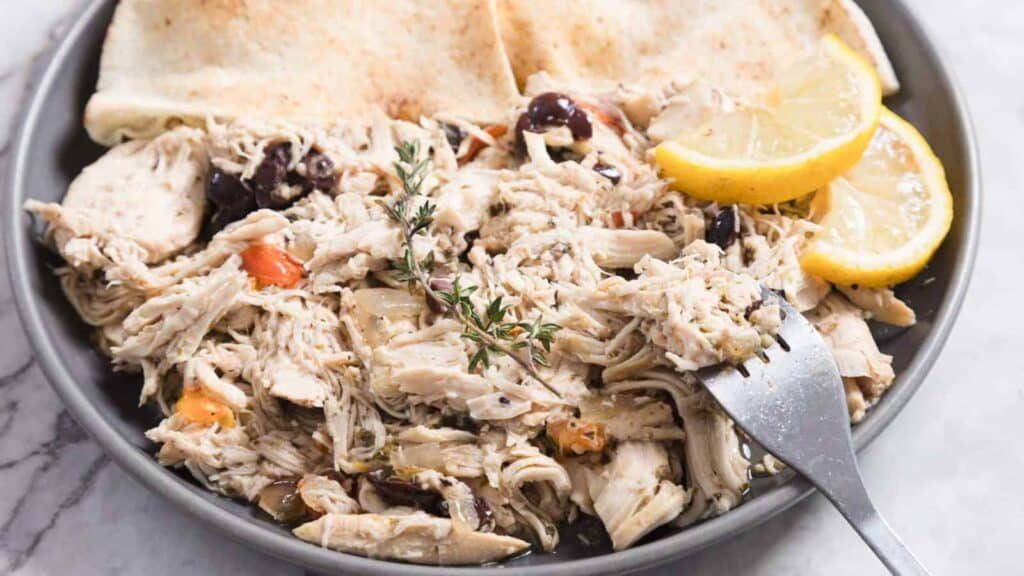 A bowl of shredded chicken mixed with olives and herbs, garnished with lemon slices and served with flatbread.