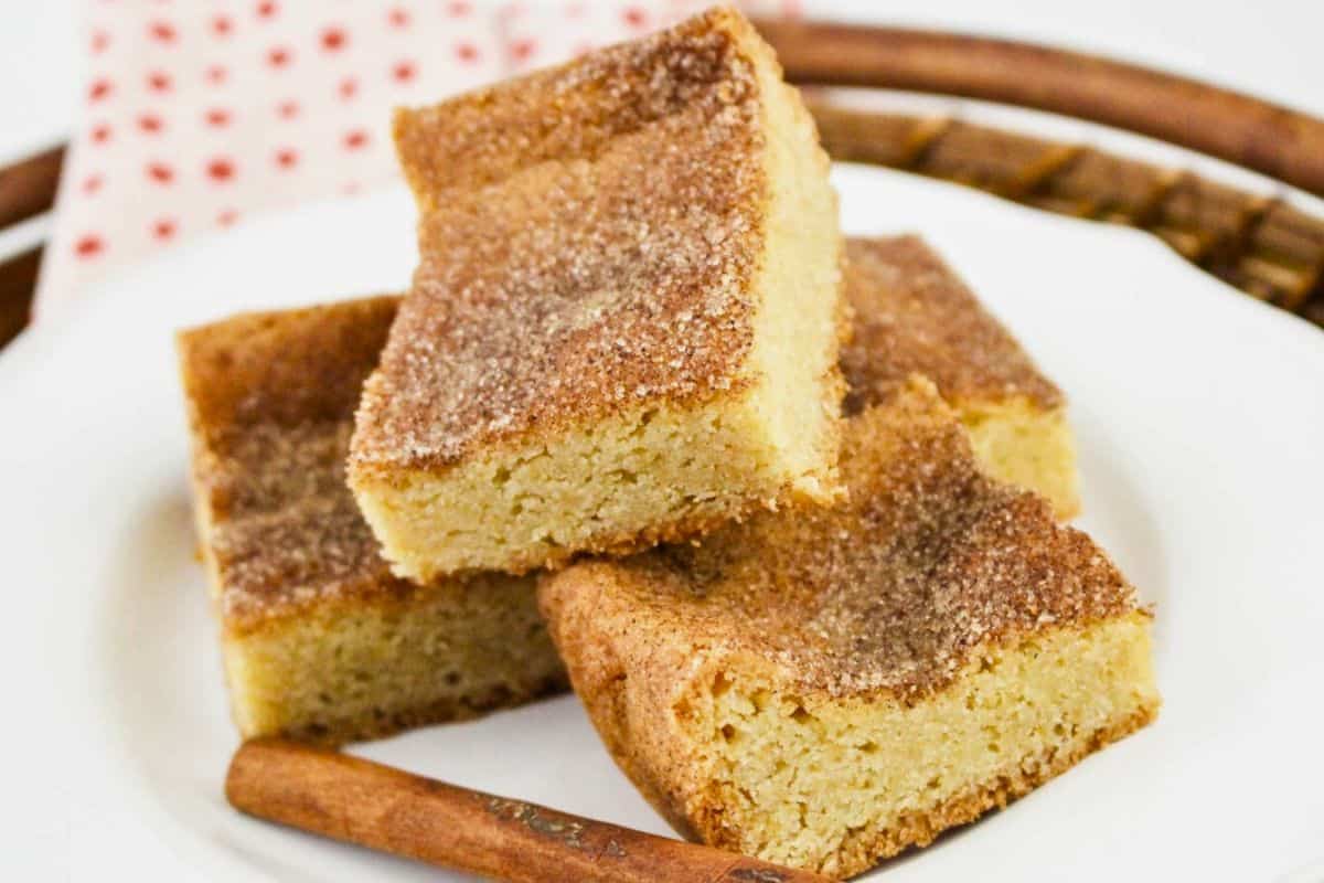 Three cinnamon sugar blondies stacked on a white plate, placed on a woven tray, with a cinnamon stick beside them.