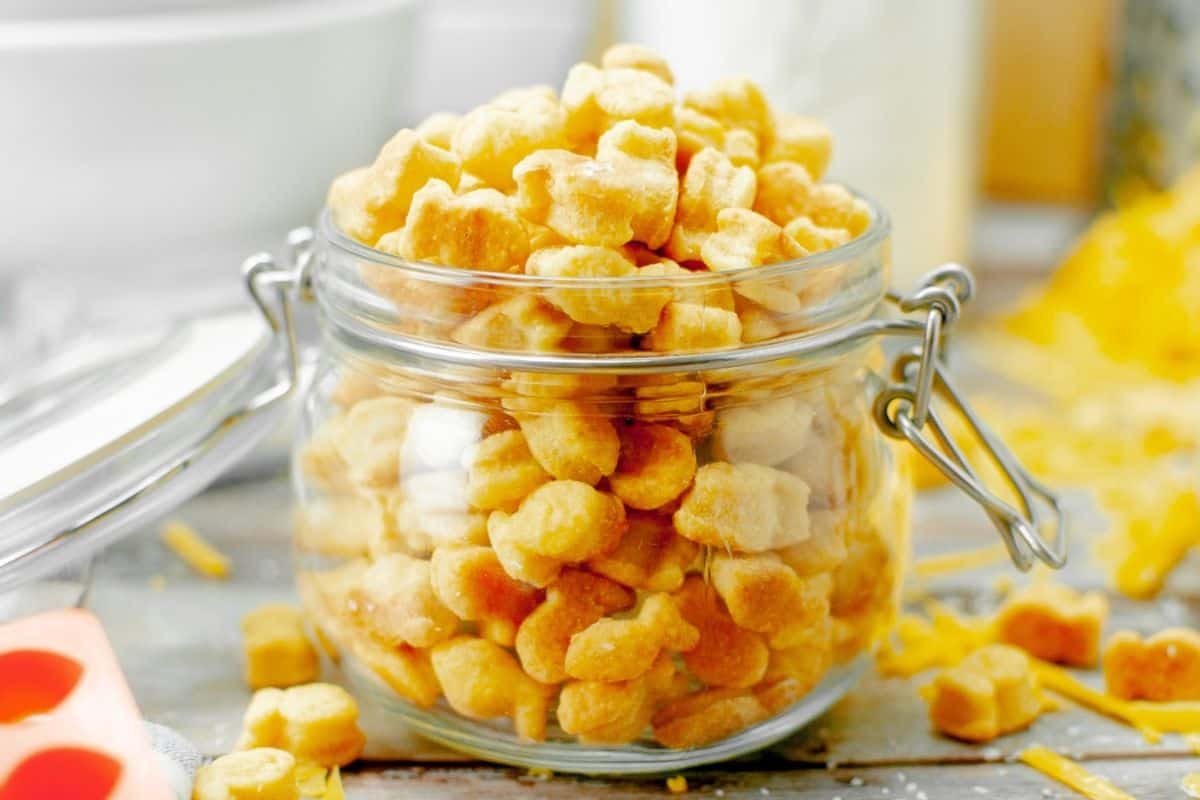 A glass jar filled with fish-shaped cheese crackers on a wooden table, surrounded by scattered crackers and cheese shreds.