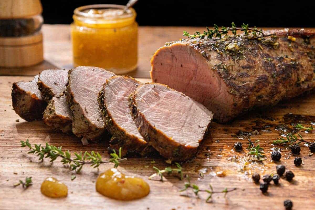 Slices of roasted pork tenderloin on a wooden board, garnished with fresh herbs. A jar of mustard is in the background, along with peppercorns scattered around the meat. The scene is warmly lit and appetizing.