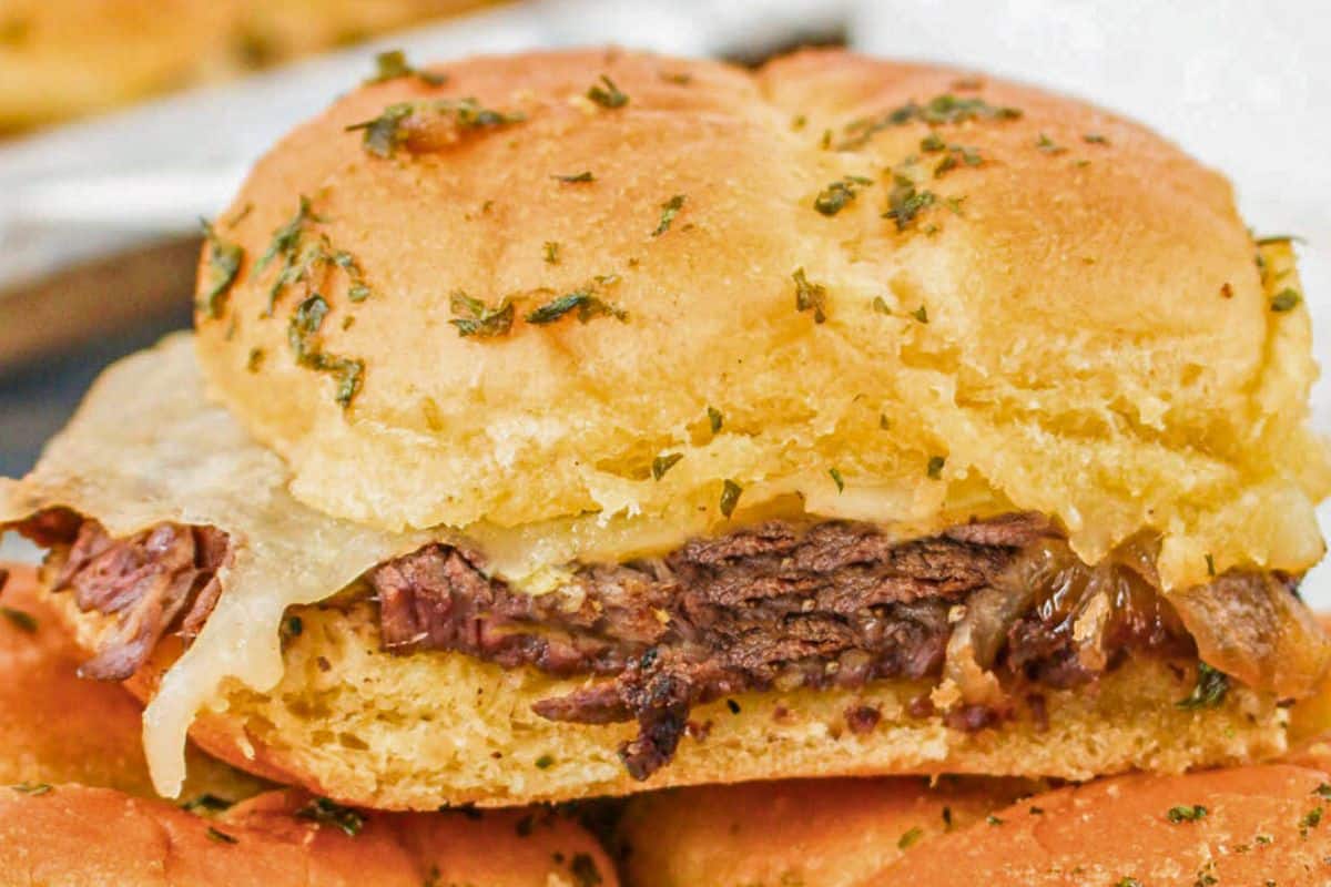 A close-up of a sandwich with shredded beef, melted cheese, and caramelized onions on a herb-topped bun.