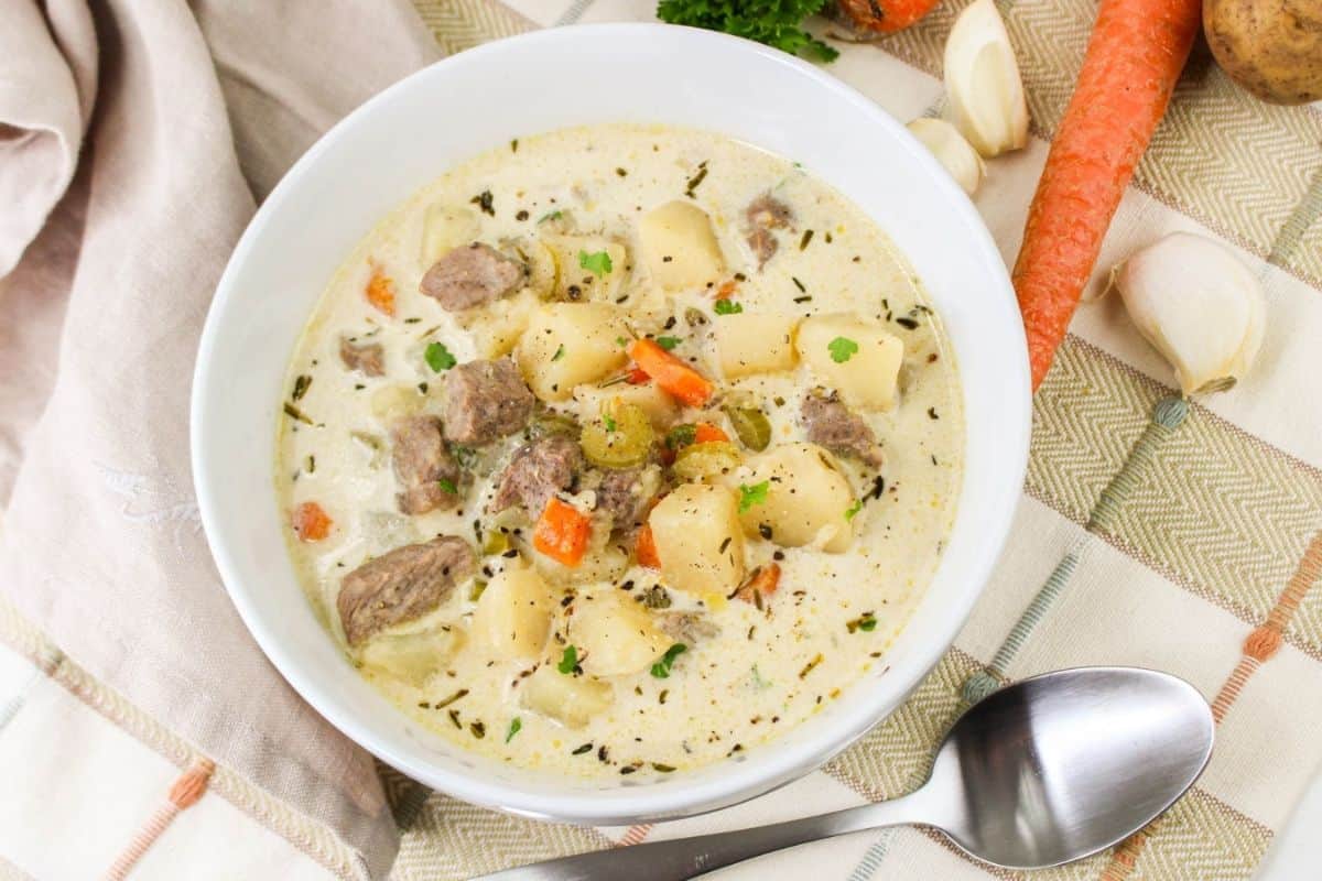 A bowl of creamy soup with chunks of meat, potatoes, and carrots, garnished with herbs. A spoon, carrot, garlic, and cloth napkin are nearby on a plaid tablecloth.