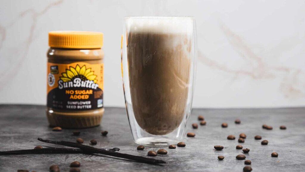 A glass of frothy iced coffee sits next to a jar of SunButter sunflower seed butter, with scattered coffee beans and a vanilla bean on a gray surface.