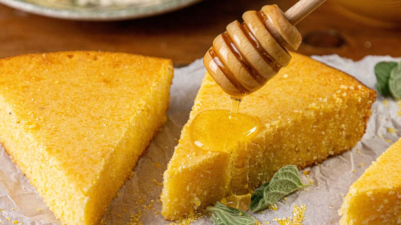 Close-up of honey being drizzled onto a slice of Southern cornbread with a wooden honey dipper. The cornbread, boasting a moist center and golden crust, sits on parchment paper, accompanied by green leaves. The softly blurred background highlights its inviting texture.