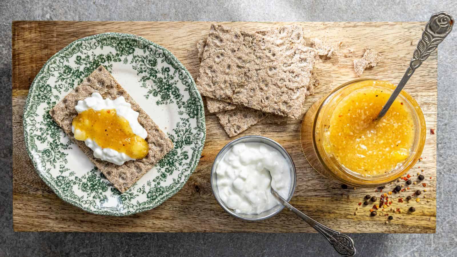 A wooden board with crispbread topped with cottage cheese and orange jam on a decorative plate. Beside it are plain crispbread pieces, a small bowl of cottage cheese, and a jar of orange jam with a spoon.