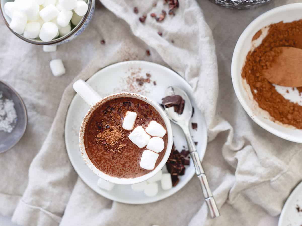 A cup of hot chocolate topped with marshmallows sits on a white plate with a spoon, surrounded by cocoa powder and extra marshmallows.