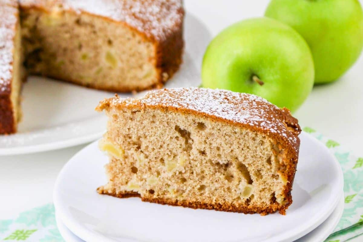 A slice of apple cake on a plate, dusted with powdered sugar. Two whole green apples are in the background, with more cake in the background on a serving plate.