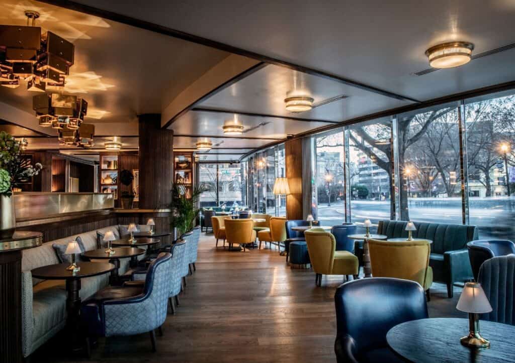 Modern restaurant interior with large windows, wooden floors, upholstered chairs, round tables, and ambient lighting, overlooking a city street with trees.