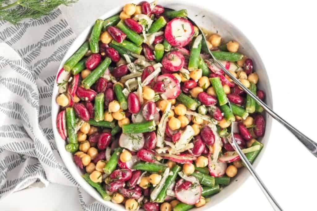 A bowl of mixed bean salad with chickpeas, red kidney beans, green beans, radishes, and herbs, served with two metal utensils.
