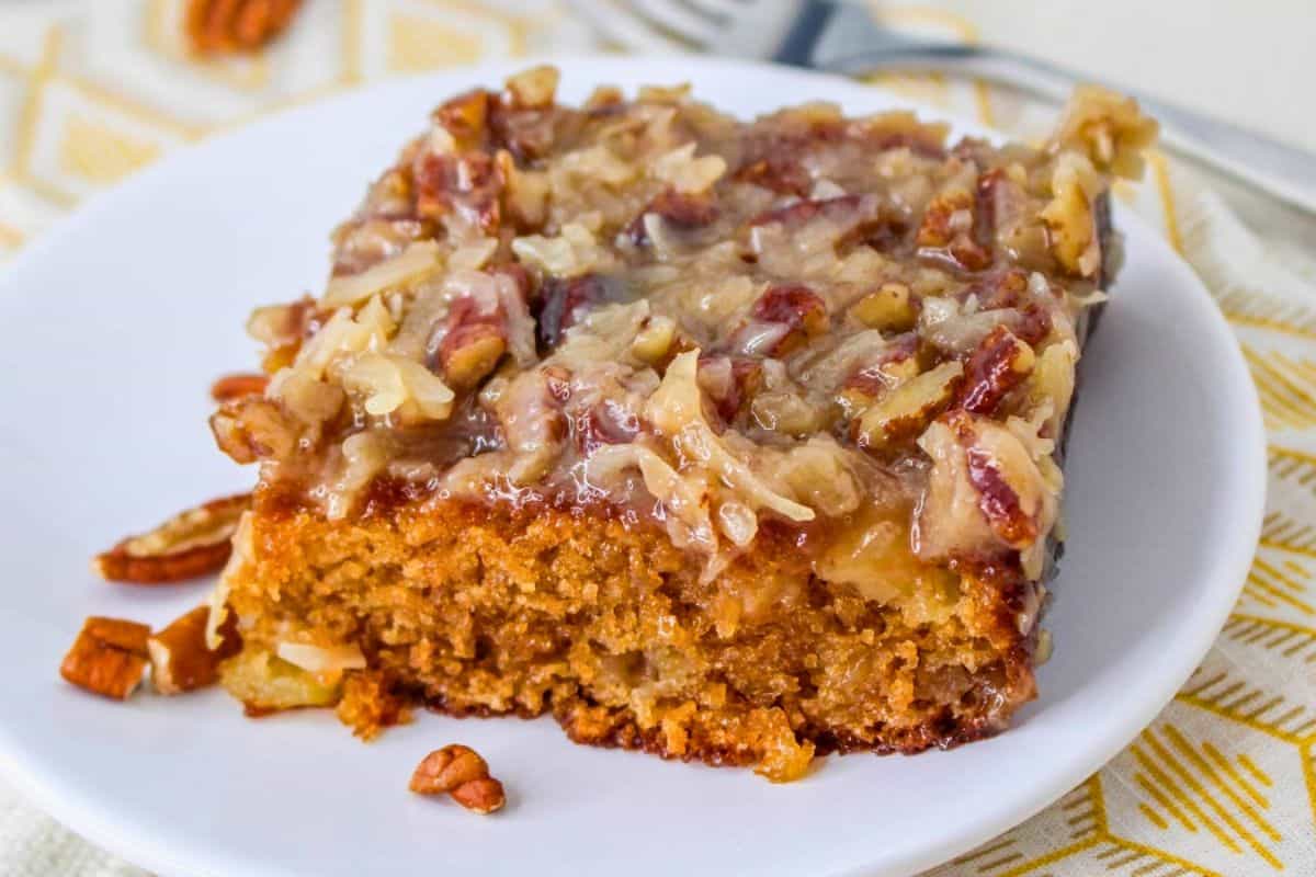 A slice of caramel pecan cake on a white plate, topped with chopped pecans and shredded coconut. A fork is in the background.
