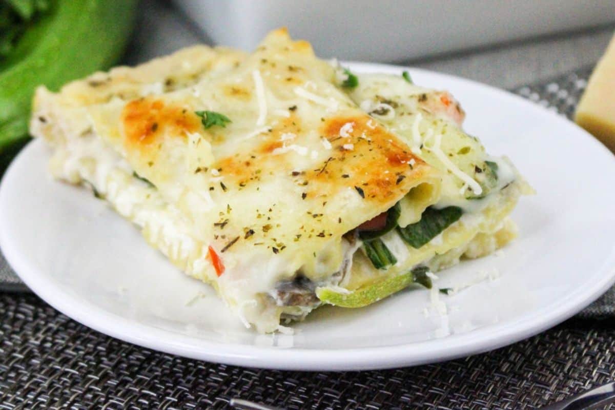 A slice of vegetable lasagna on a white plate, featuring layers of pasta, cheese, and vegetables, garnished with herbs and grated cheese.