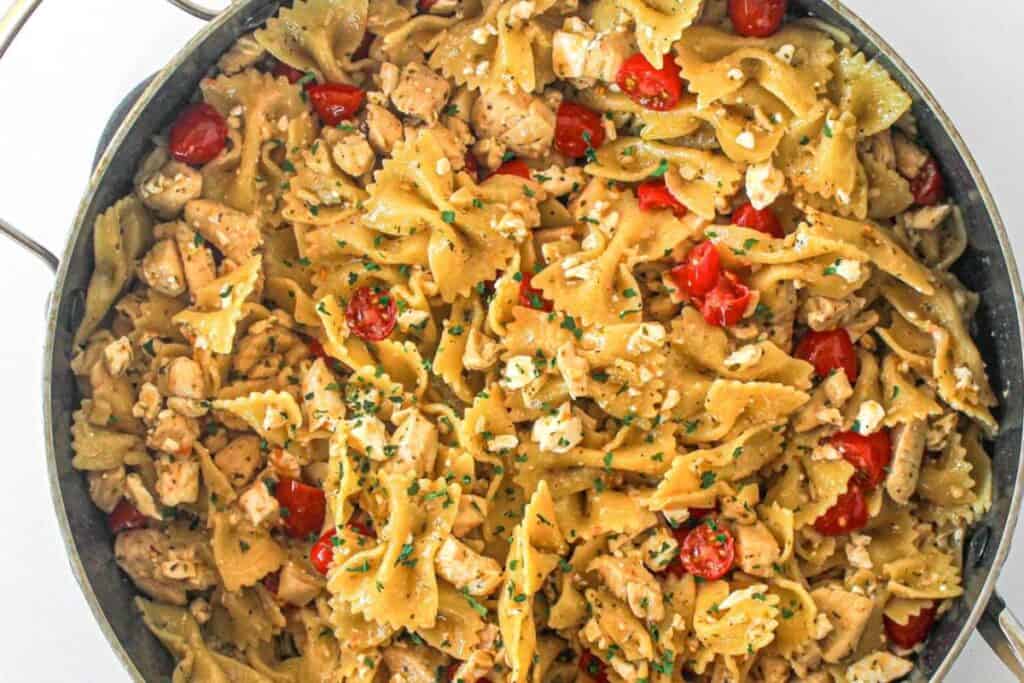 A pan filled with bowtie pasta mixed with diced chicken, cherry tomatoes, feta cheese, and chopped herbs.