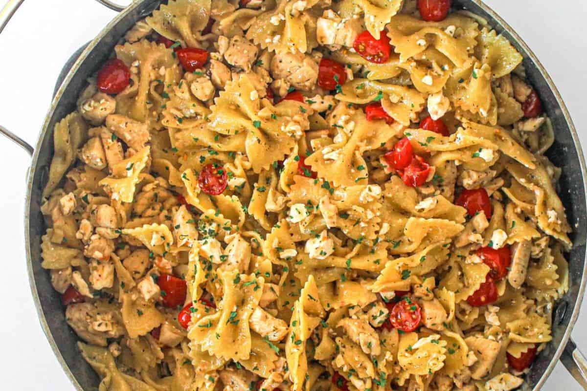 A pan filled with bowtie pasta mixed with diced chicken, cherry tomatoes, feta cheese, and chopped herbs.