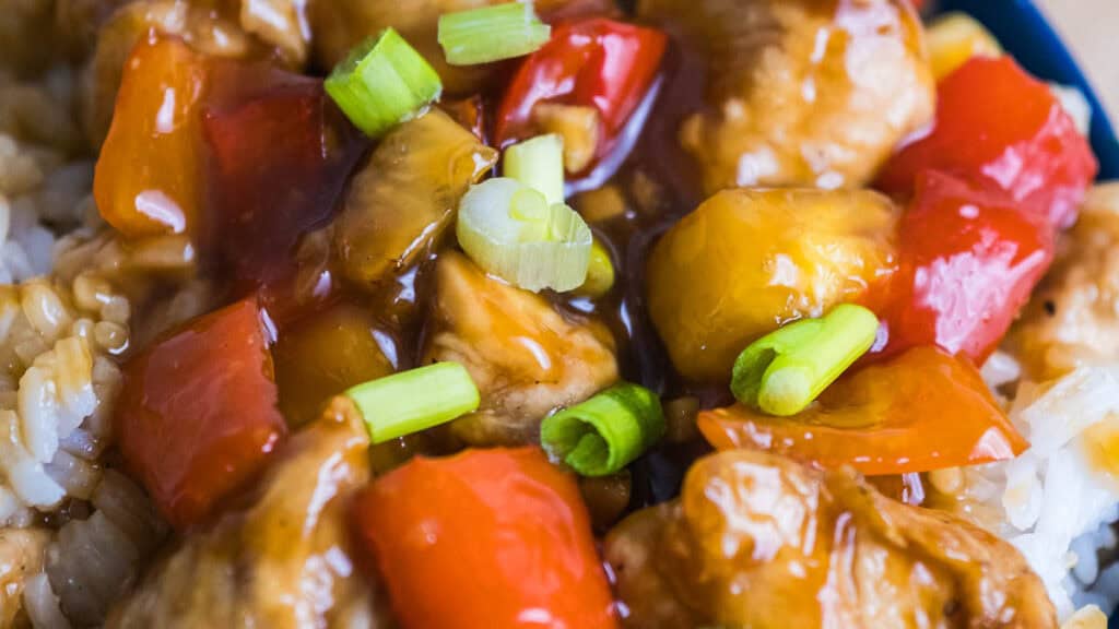 Close-up of sweet and sour chicken with red and yellow bell peppers, green onions, and sauce served over white rice.