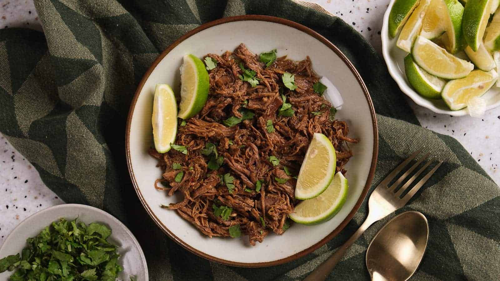 A plate of beef barbacoa garnished with chopped cilantro and lime wedges, placed on a green cloth.
