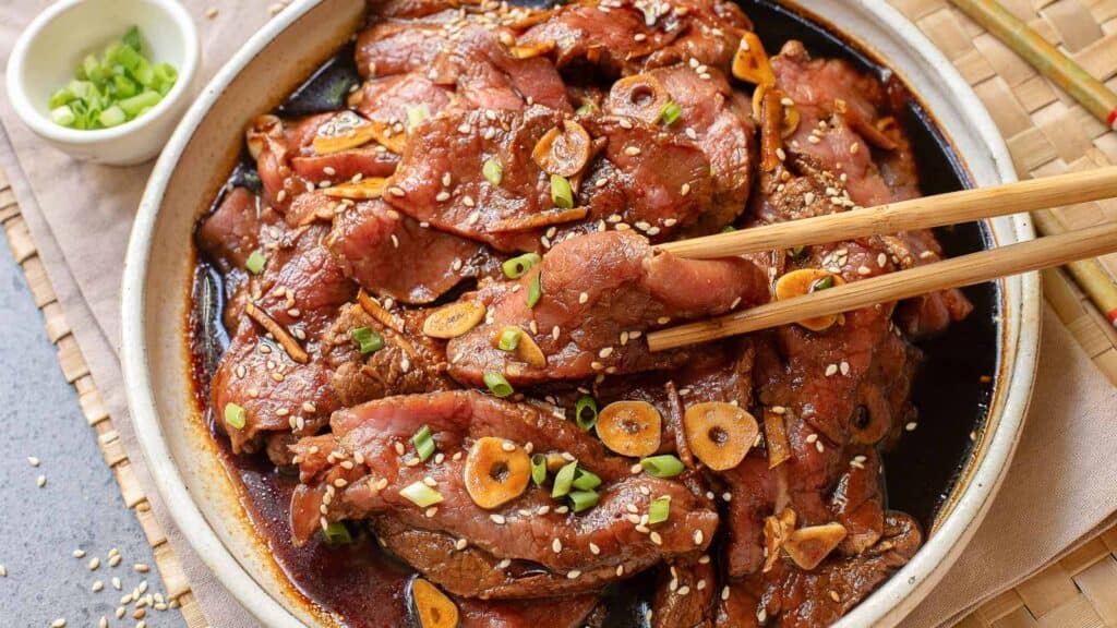 A bowl of marinated beef slices garnished with sesame seeds, sliced garlic, and chopped green onions, with chopsticks picking up a piece.