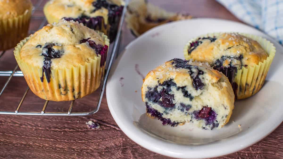 Blueberry muffins on a cooling rack next to a white plate with one muffin, partially eaten.