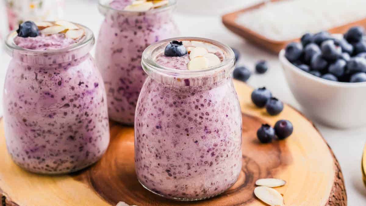 Three glass jars filled with blueberry chia pudding, topped with sliced almonds and blueberries, placed on a wooden board with a bowl of blueberries in the background.