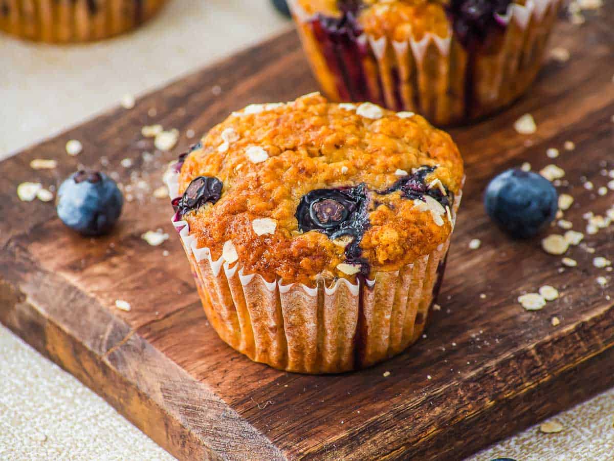 A blueberry muffin in a paper liner sits on a wooden board with fresh blueberries and oats scattered around.