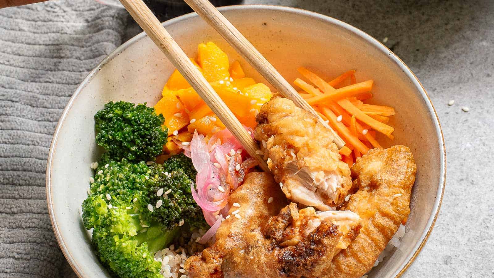 Bowl with fried chicken, sliced carrots, broccoli, pickled onions, and mango over rice, sprinkled with sesame seeds. Chopsticks holding a piece of chicken.