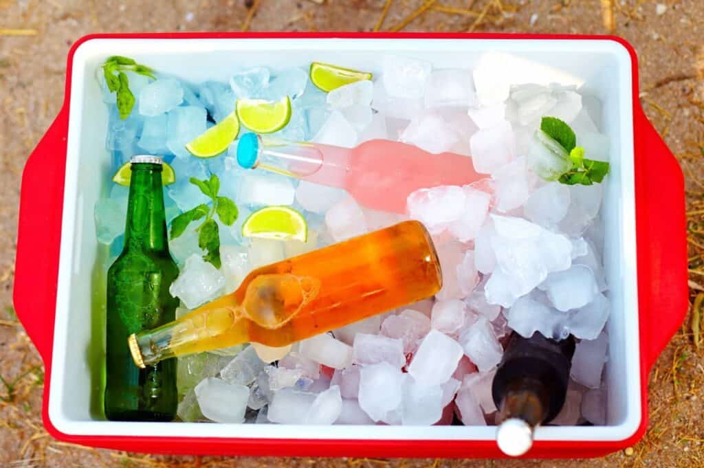 A red cooler filled with ice holds bottles of drinks, lime wedges, and mint sprigs, set on sandy ground.