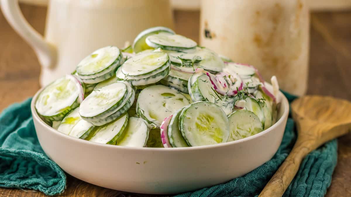 A bowl of cucumber salad with dill and red onions, dressed in a creamy sauce, on a wooden table beside a wooden spoon and jars.