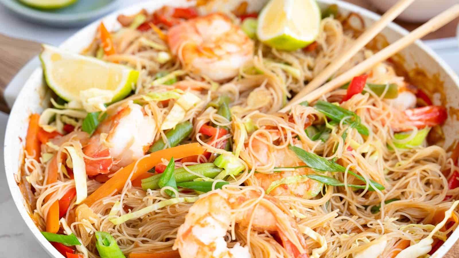 A close-up of a plate of stir-fried rice noodles with shrimp, sliced bell peppers, cabbage, green onions, and lime wedges. Chopsticks rest on top of the colorful, vibrant dish.