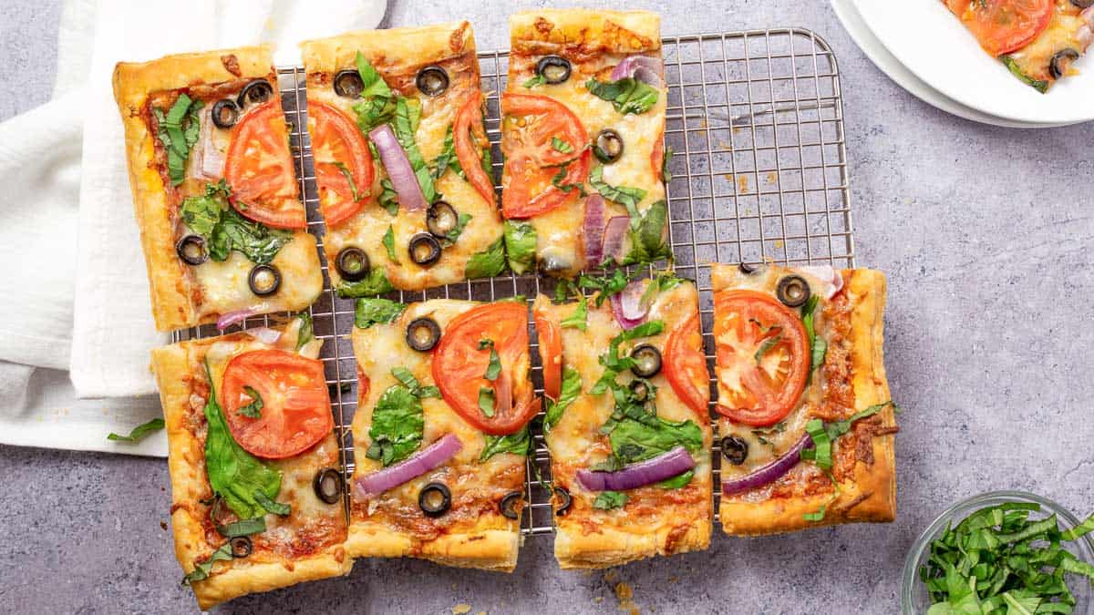 Six rectangular vegetable pizzas with tomatoes, spinach, olives, and red onions on a cooling rack, with a white napkin nearby.