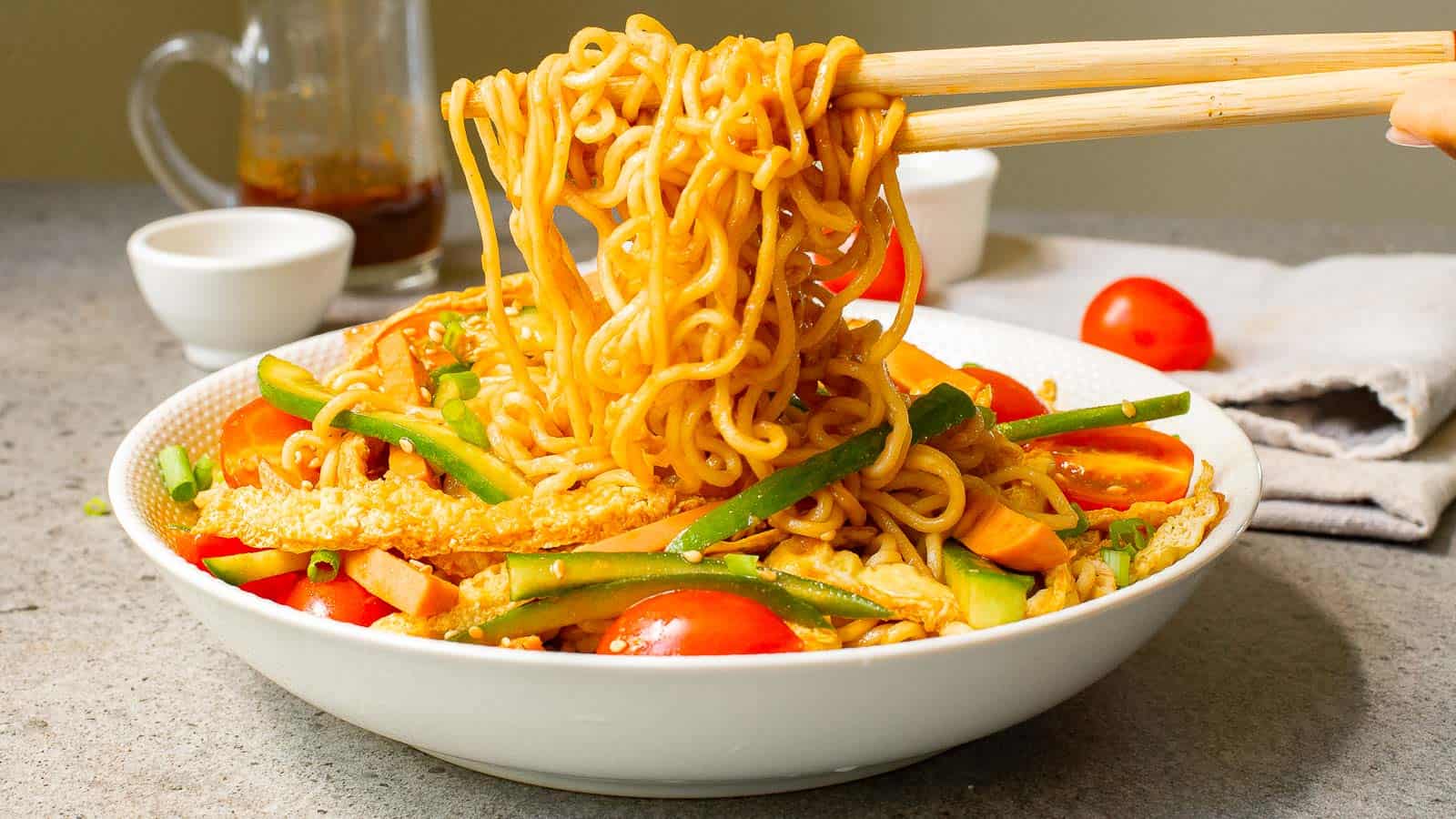 A bowl of stir-fried noodles with chopsticks lifting a portion. The dish includes sliced cucumbers, tomatoes, and green onions. A sauce jug and small bowl are in the background. The scene is set on a stone countertop.