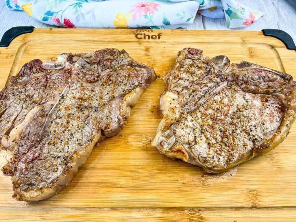 Two cooked T-bone steaks seasoned with pepper and spices are placed on a wooden cutting board with a floral cloth in the background.