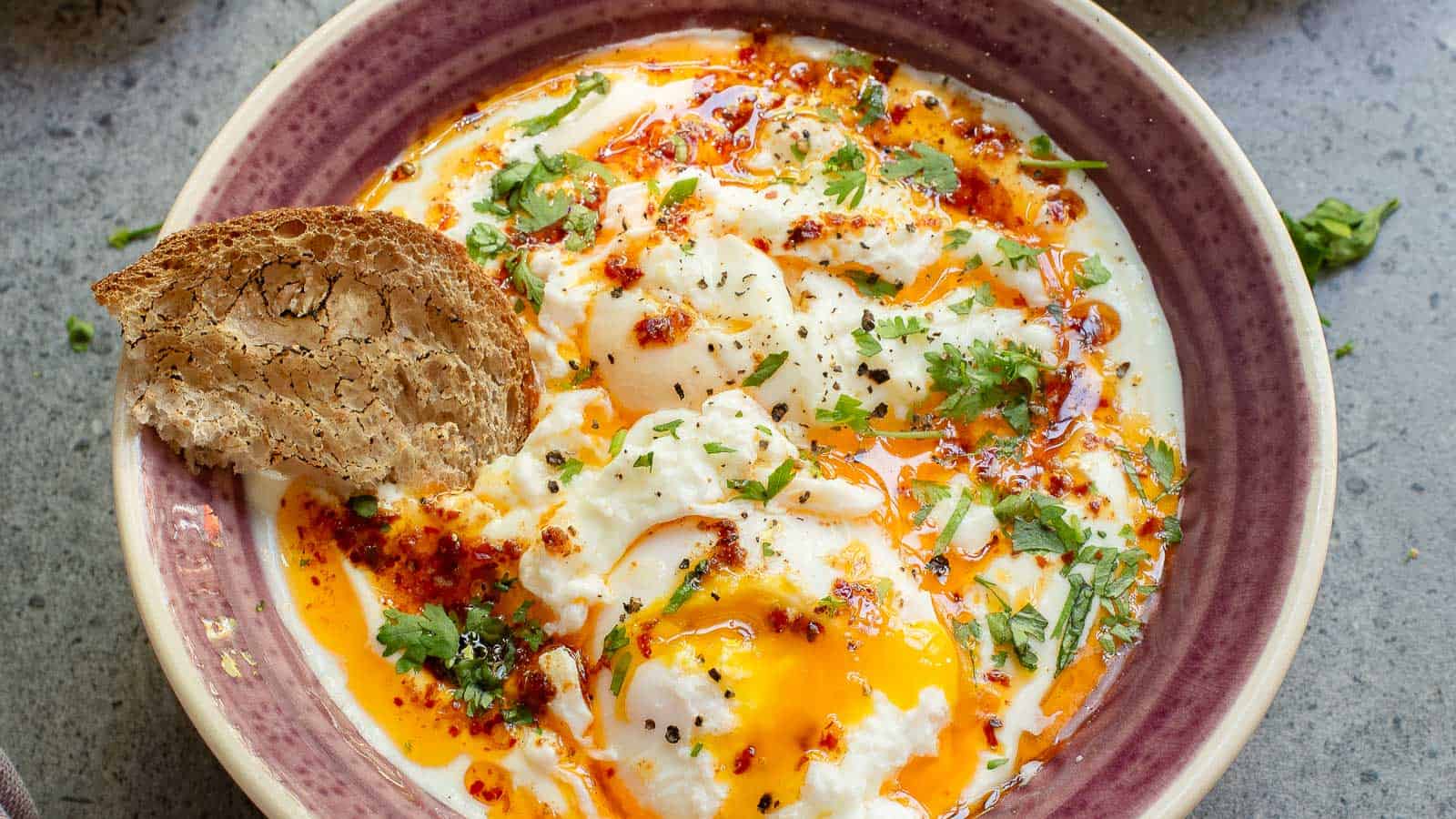 A bowl of Turkish Eggs: poached eggs over garlic yogurt, topped with herbs and chili butter, served with a slice of bread on the side.