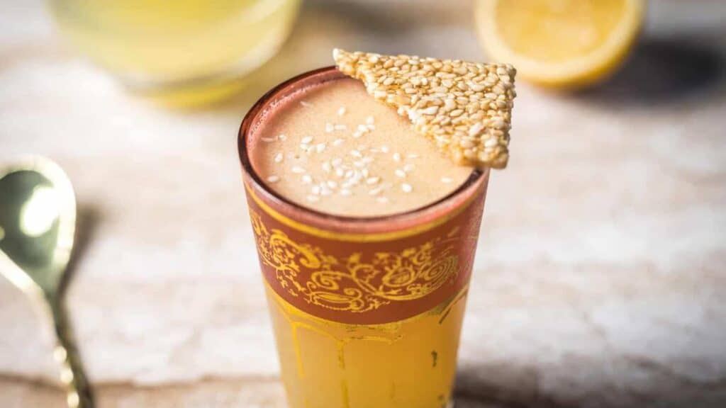 A tall glass with a tan frothy drink, topped with sesame seeds and a sesame brittle, sits on a light surface with a gold spoon and lemon half nearby.