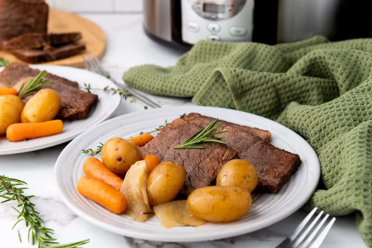 A plate of pot roast with sliced beef, baby potatoes, carrots, and onions, garnished with rosemary, sits on a table next to a green towel and a slow cooker.