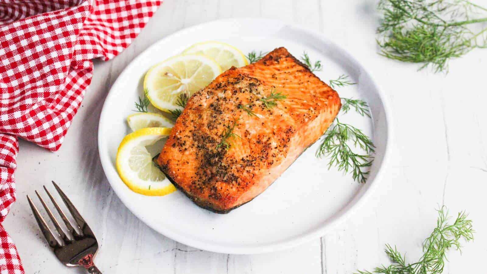 Grilled salmon fillet on a white plate garnished with lemon slices and dill, next to a red checkered cloth and a fork.