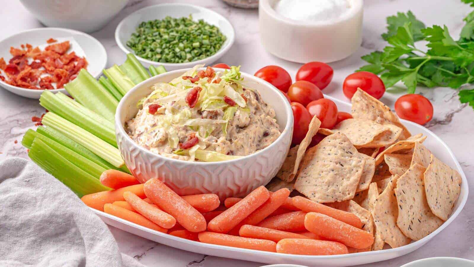 A platter with a bowl of dip topped with bacon bits, surrounded by celery sticks, baby carrots, cherry tomatoes, and tortilla chips.