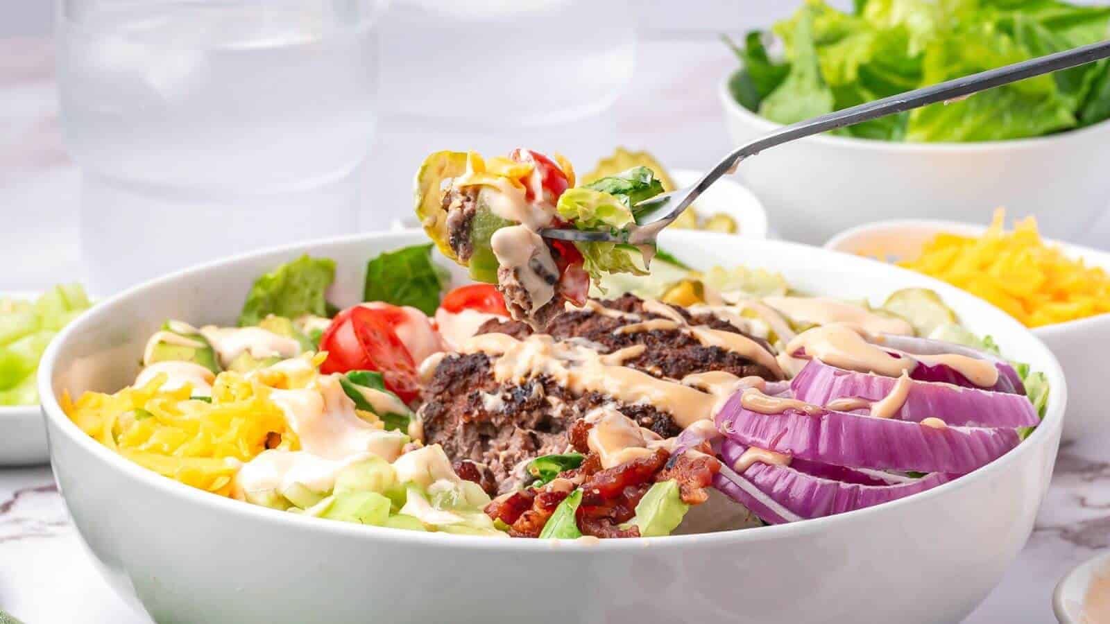 A bowl of salad with sliced red onions, lettuce, tomatoes, shredded cheese, ground beef, bacon, and dressing; a fork holds a bite above the bowl.