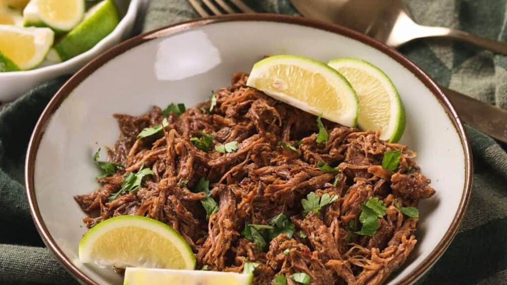 A plate of beef barbacoa garnished with chopped cilantro and lime wedges, placed on a green cloth.