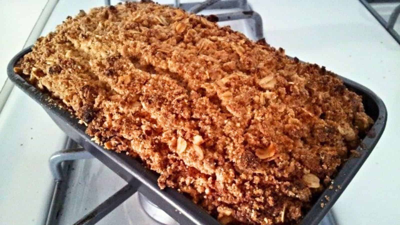 Image shows a loaf of baked bread with a golden brown, crumbly oat topping sits in a metal pan on a stove.