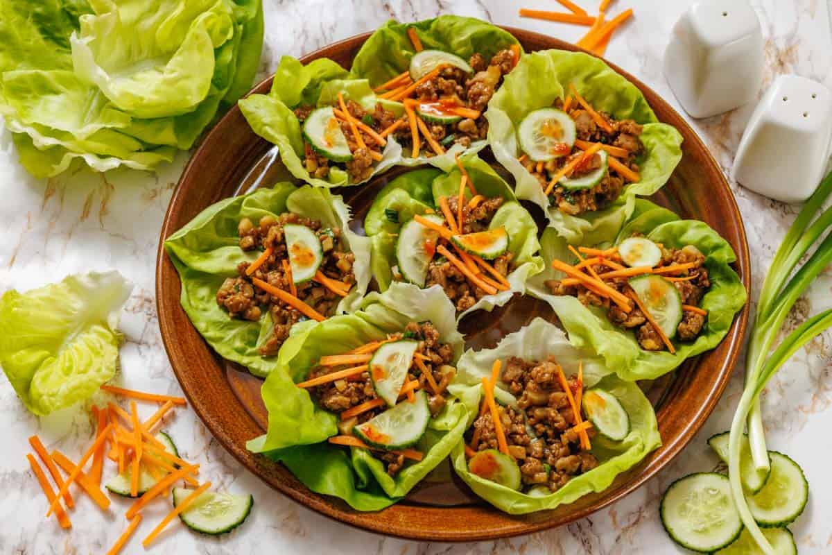A wooden plate with Chicken lettuce wraps surrounded by extra lettuce leaves and vegetables on a marble surface.