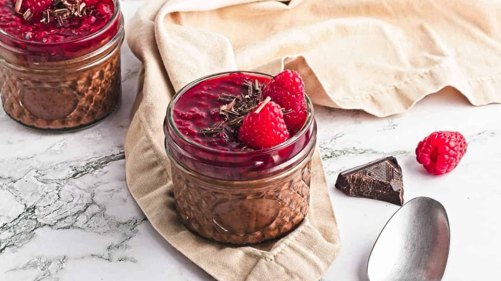 Two small jars filled with chocolate mousse, topped with raspberry sauce, fresh raspberries, and chocolate shavings. A beige cloth, chocolate pieces, loose raspberries, and a spoon are nearby on a marble surface.