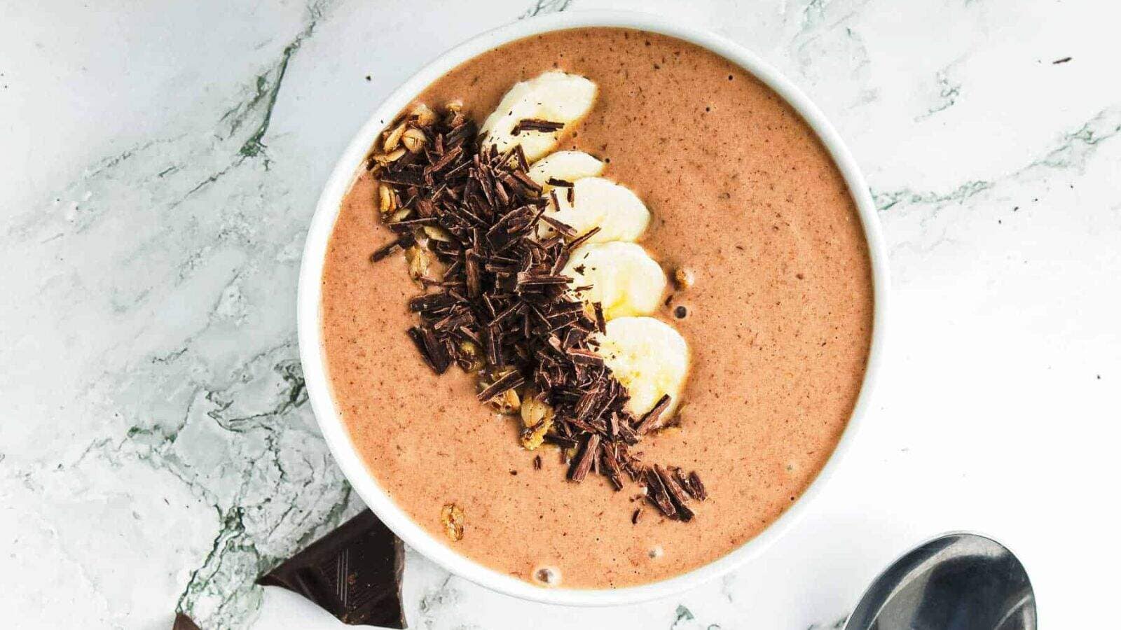 A bowl filled with a chocolate smoothie, topped with banana slices, granola, and chocolate shavings, sits on a white marble surface next to a spoon and a piece of chocolate.