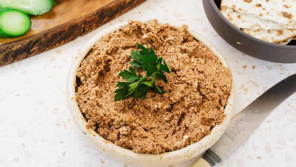 A bowl of chopped liver garnished with parsley sits on a countertop next to a knife, matzo crackers, and sliced cucumbers.