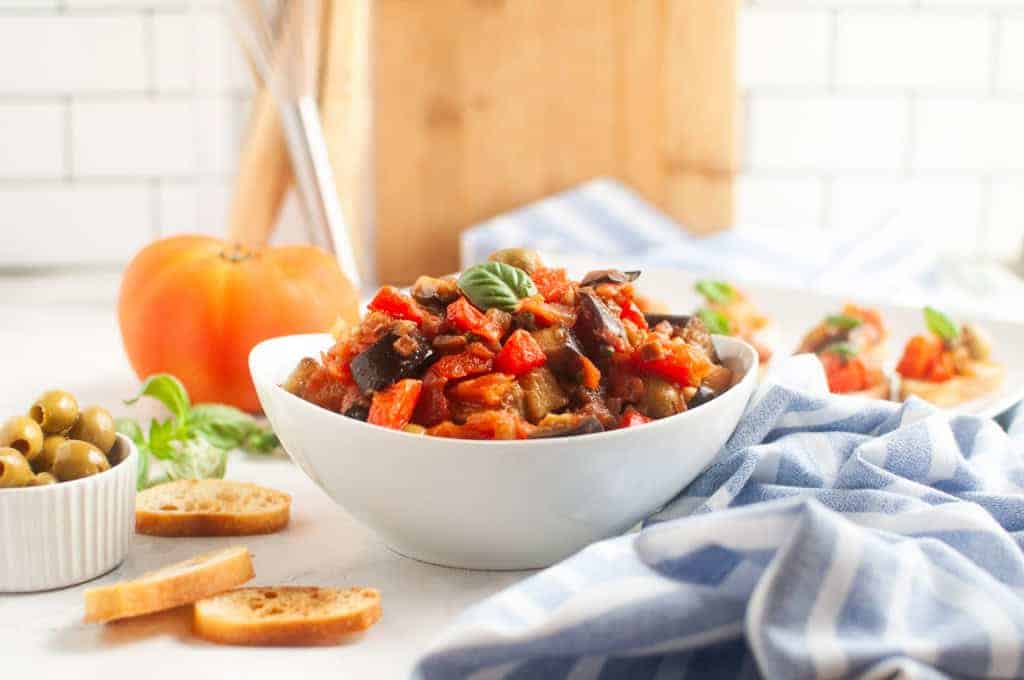 A white bowl filled with Low Carb Caponata, garnished with a basil leaf, sits on a table with bread slices, olives, and a blue-striped cloth nearby.