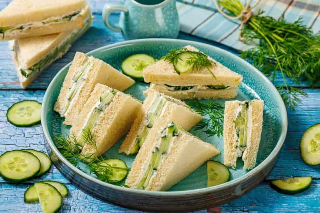 A plate of cucumber sandwiches on white bread, garnished with fresh dill and cucumber slices, sits on a blue wooden table with a jar and herbs in the background.