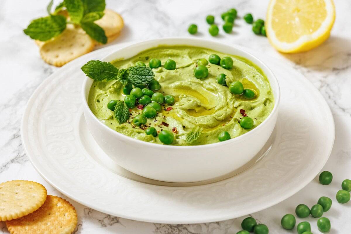 A white bowl of vibrant green Green Pea Hummus, garnished with peas and mint, served with crackers and a lemon wedge.