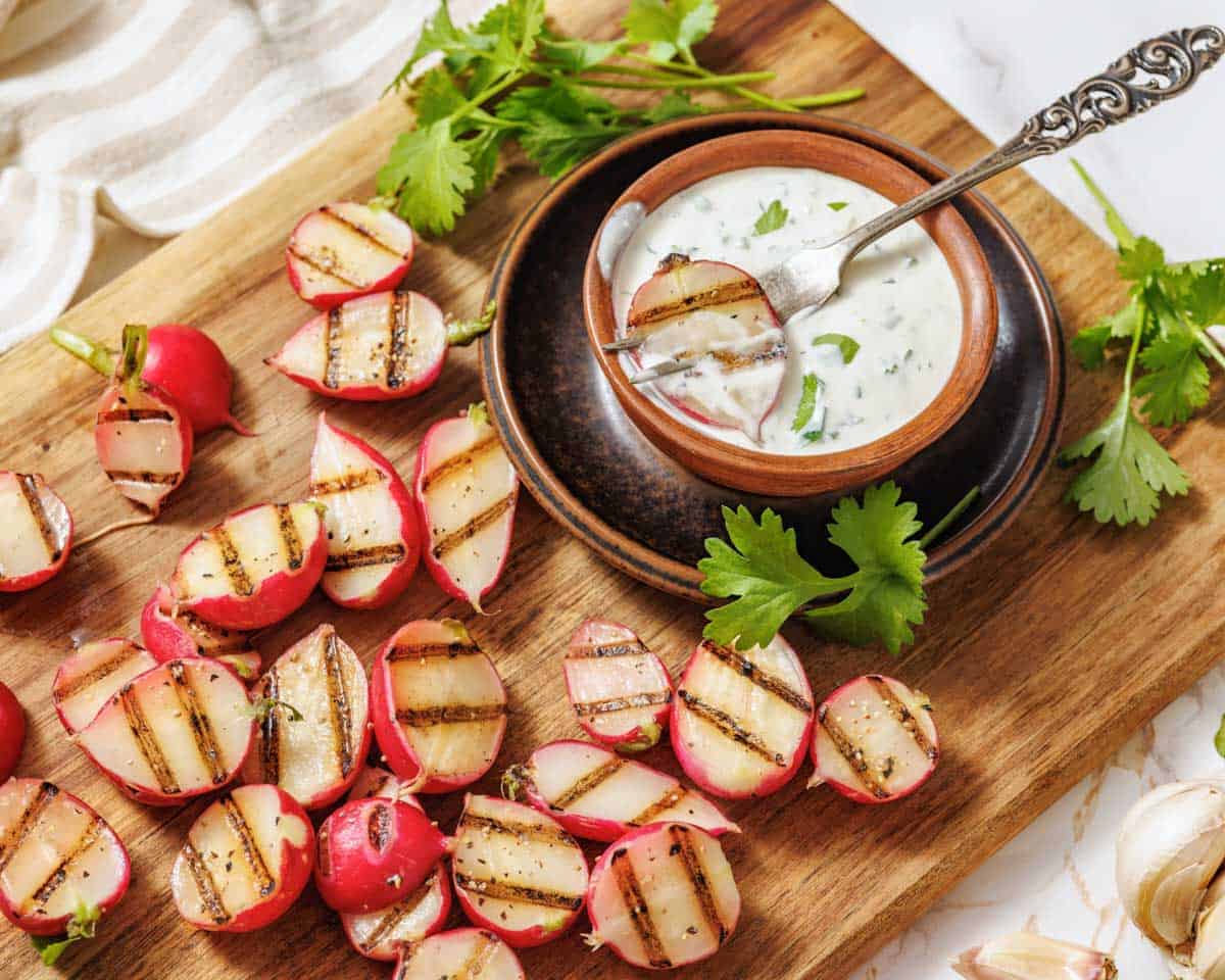 Grilled radish halves with char marks served on a rustic wooden board, accompanied by a small bowl of creamy jalapeño dip garnished with cilantro and a decorative spoon, surrounded by fresh herbs and garlic cloves.